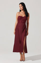 A woman is wearing a sleeveless, burgundy dress with spaghetti straps. The dress has a fitted, lace bodice with a zipper down the center and a long, flowing skirt that features a high slit on one side. She is also wearing strappy gold high-heeled sandals.